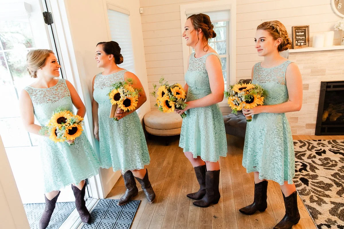 Four bridesmaids in matching teal lace dresses and brown boots hold sunflower bouquets, standing cheerfully in a bright room with wooden floors.