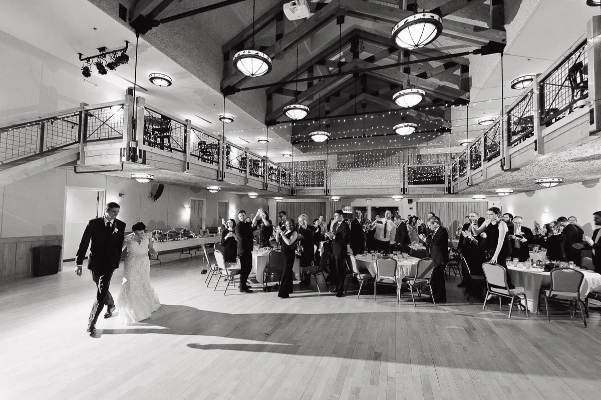 A newlywed couple joyfully enters a grand Silverthorne Pavilion reception hall, guests stand applauding. Elegant decor with string lights and high ceilings creates a festive atmosphere.