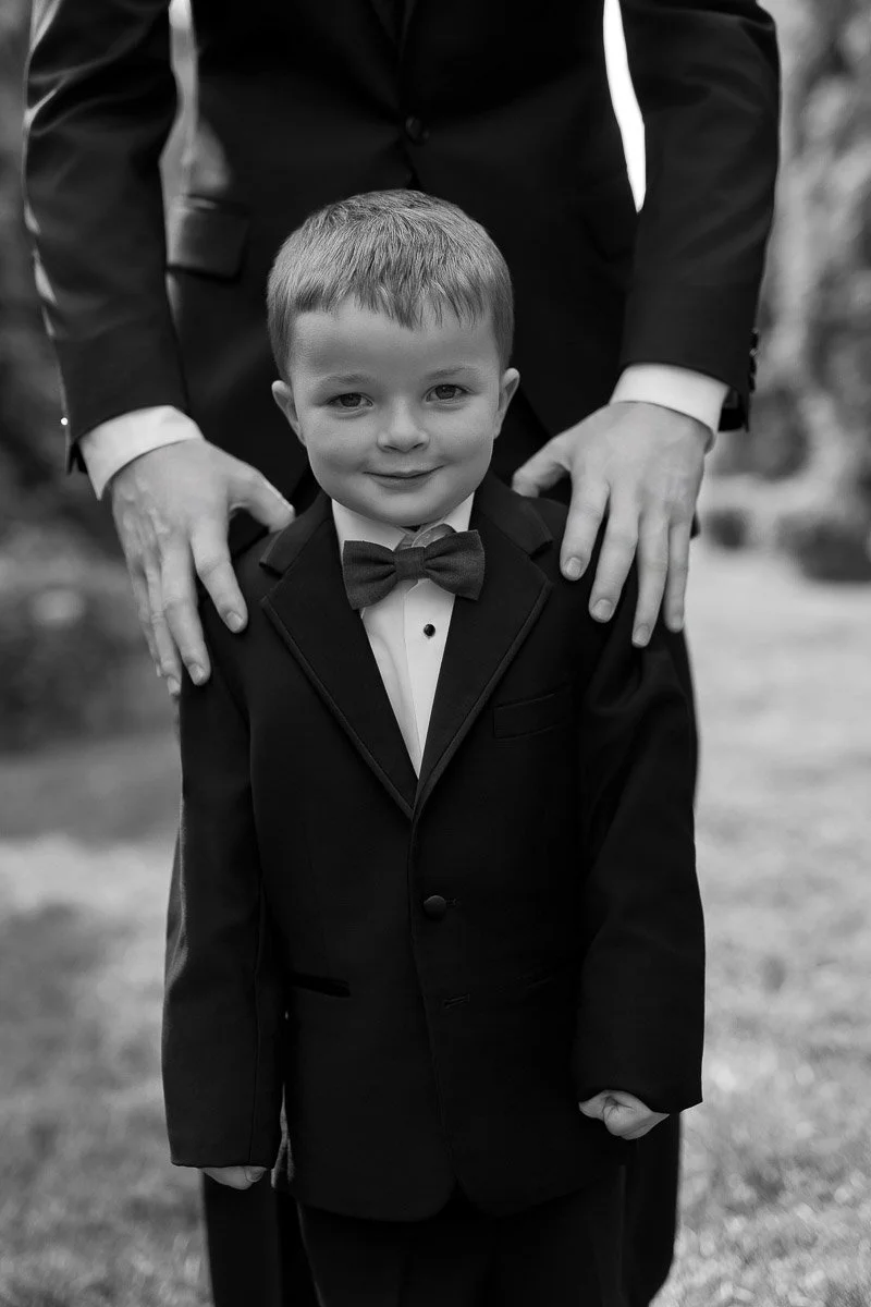 Young boy in a tuxedo with a bowtie stands outdoors, smiling warmly. Adult hands rest on his shoulders, conveying a sense of care and protection.