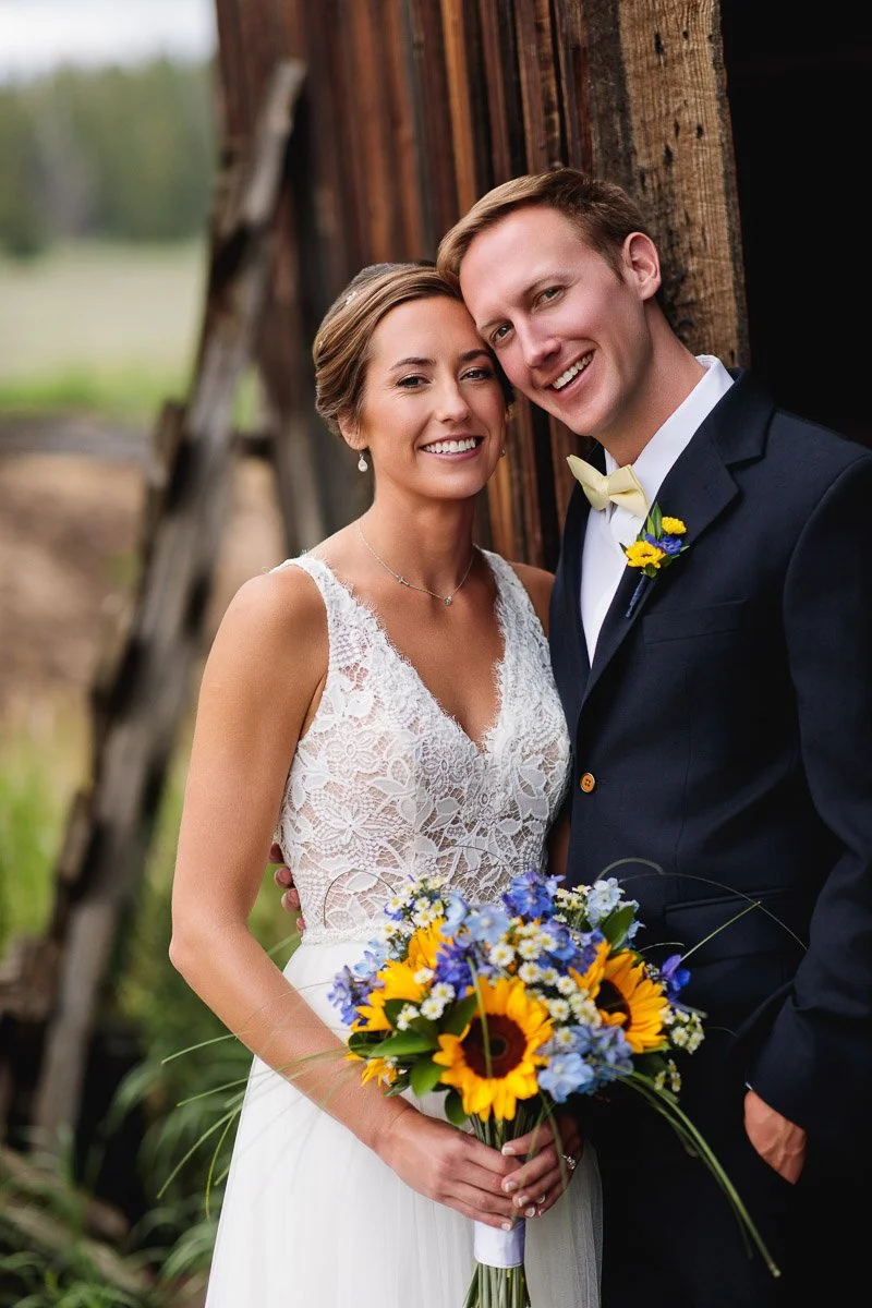 A joyful couple in wedding attire poses by a rustic wooden wall. The bride holds a bouquet of sunflowers and blue flowers, conveying a warm and cheerful tone.