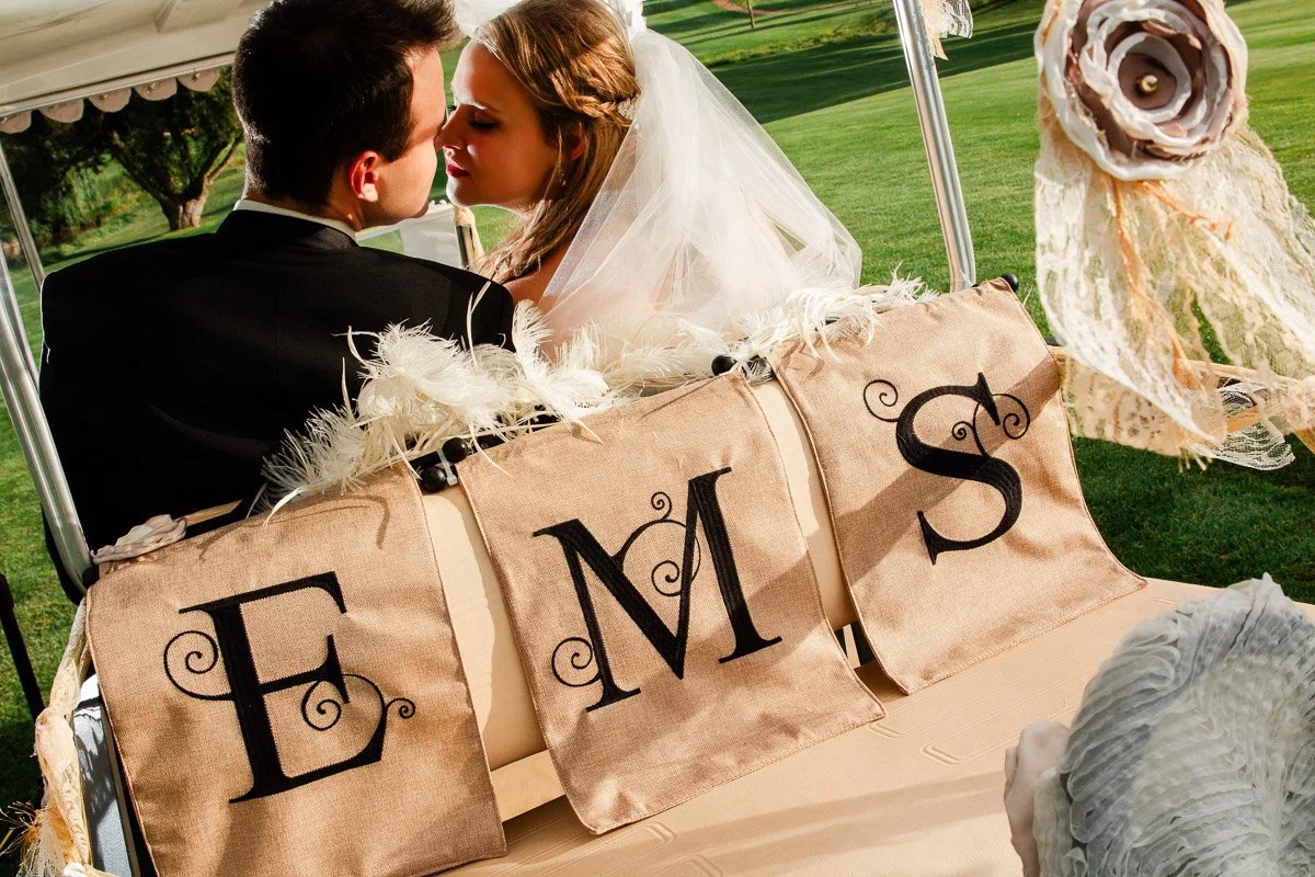 A bride and groom share a romantic moment on a decorative golf cart with burlap banners reading "EMS." The scene conveys love and celebration.