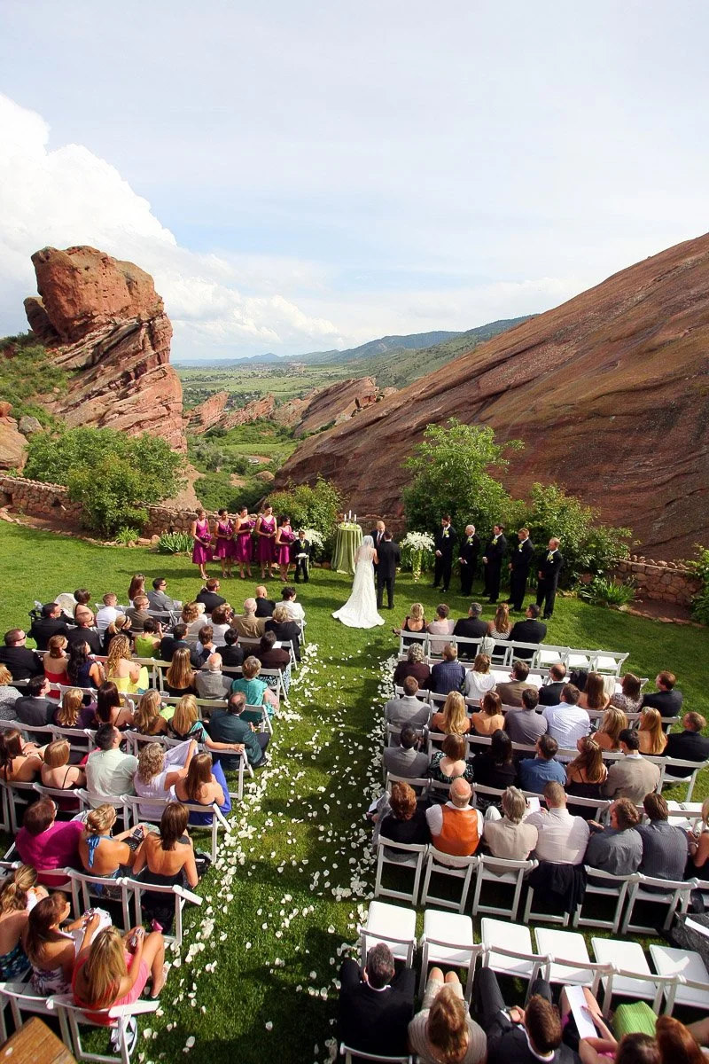 A wedding ceremony is set outdoors at Red Rocks Amphitheatre. Guests sit on white chairs, framing an aisle of rose petals. Bridal party stands against a backdrop of towering red rock formations and scenic green landscape under a clear sky.