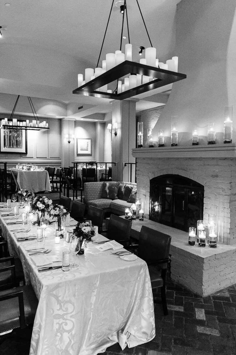 Elegant dining room in black and white, featuring a long table set for guests, a glowing fireplace with candles, and chic chandeliers creating a warm ambiance.