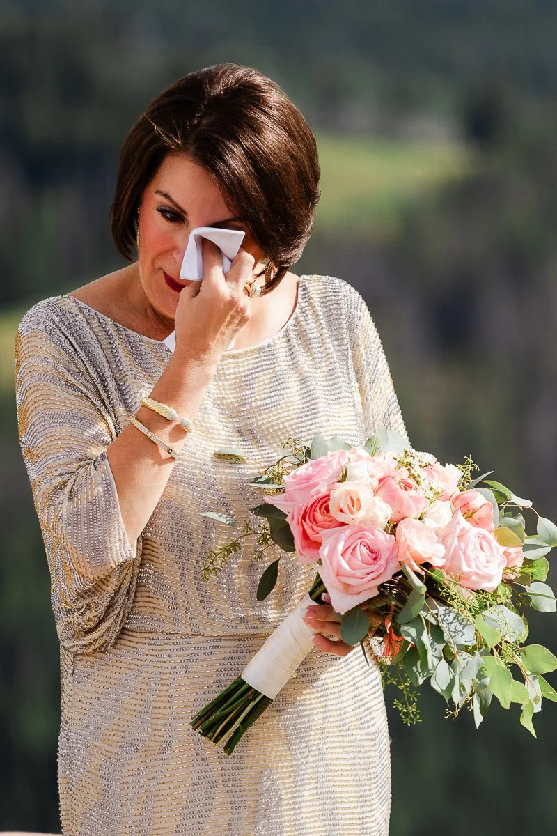 A woman in a shimmering dress wipes a tear with a tissue, holding a bouquet of pink roses and greenery. The setting is serene and emotional.