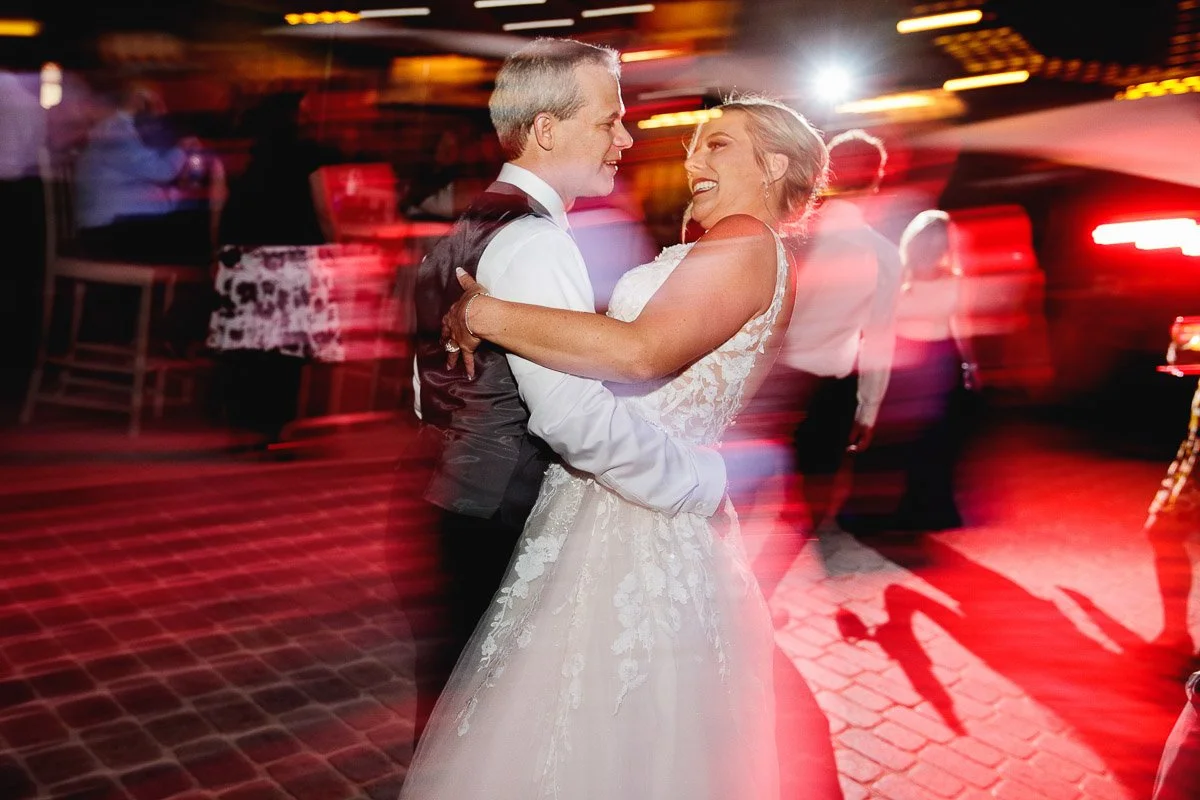 A joyful couple dances closely at a wedding reception, with colorful motion blur and bright lights creating a festive, lively atmosphere.