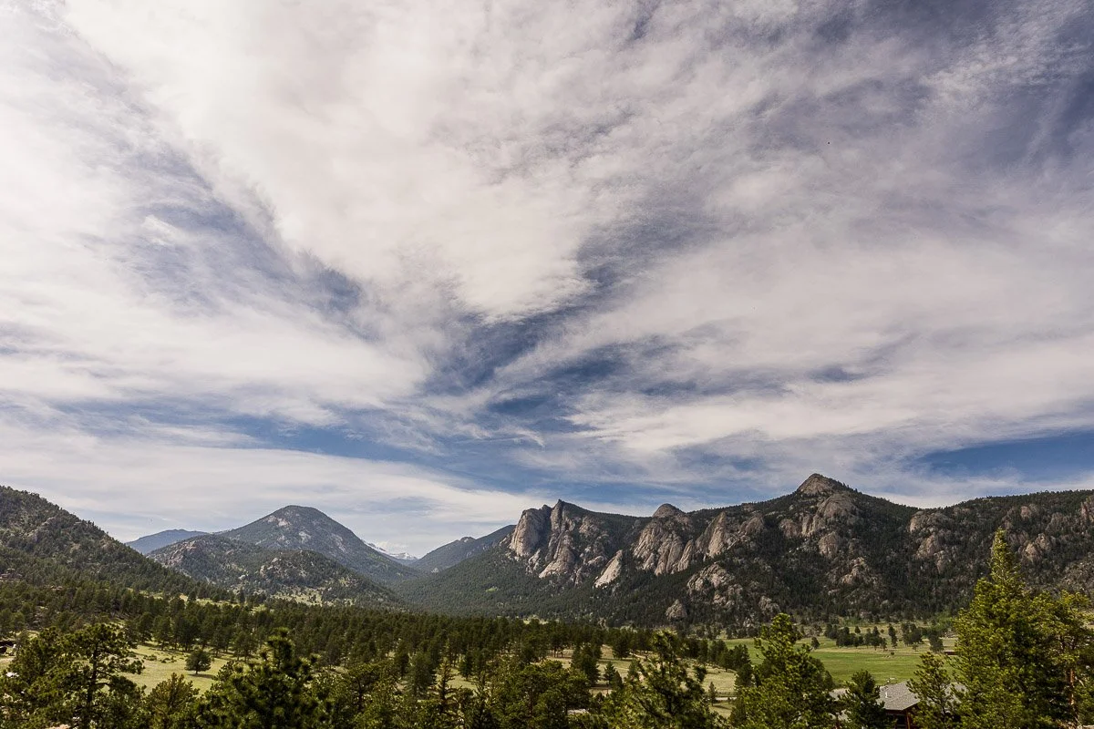 Wide landscape view of rocky mountains under a dramatic sky with scattered clouds. Foreground features green forest. Calm, expansive atmosphere.