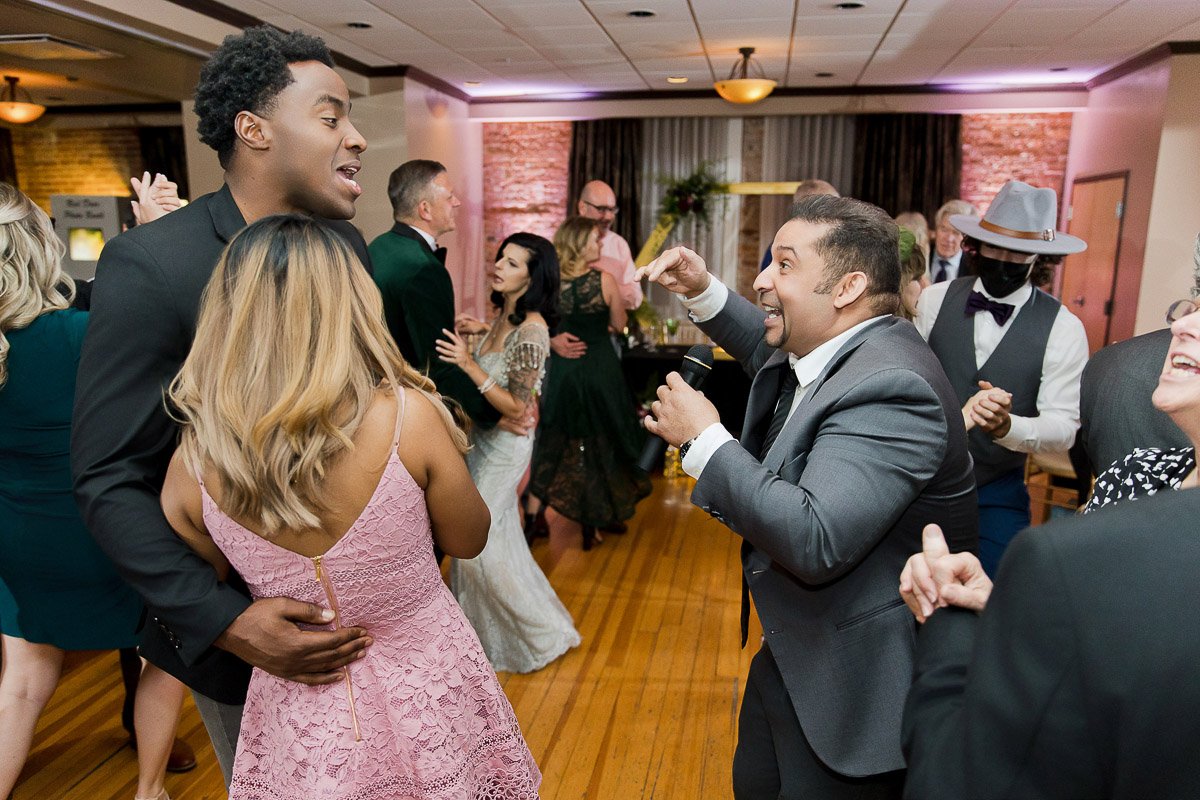 A lively wedding reception with guests dancing and laughing. A man in a gray suit passionately sings into a microphone, while couples dance joyfully.