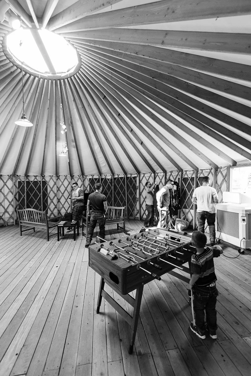 Black and white photo of people inside a spacious yurt with a wooden floor and circular roof window. A child plays foosball, adults stand nearby. Relaxed atmosphere.