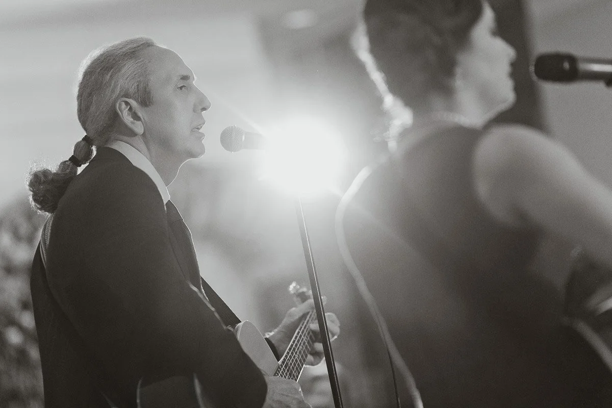 Black and white image of two musicians singing and playing guitars on stage, illuminated by bright stage light, creating a warm, intimate atmosphere.