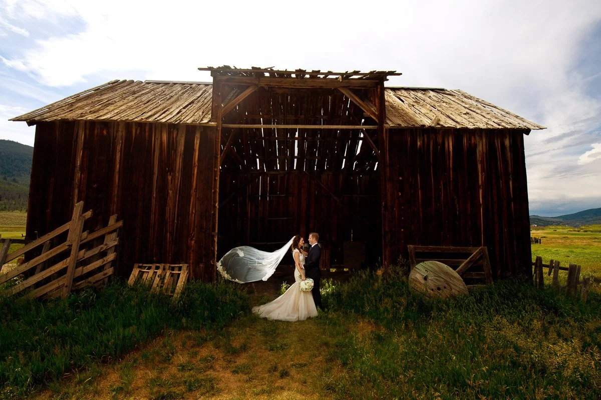 A couple in wedding attire stands in front of an old wooden barn. The bride's veil flows dramatically, and lush fields extend in the background by Keystone wedding photographer tomKphoto.