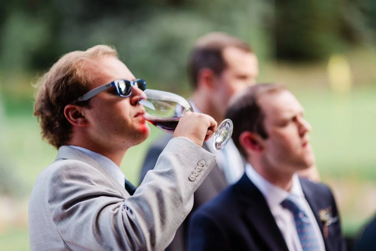 A man in sunglasses and a light gray suit sips wine from a glass, with two others in suits blurred in the background, outdoors in daylight.