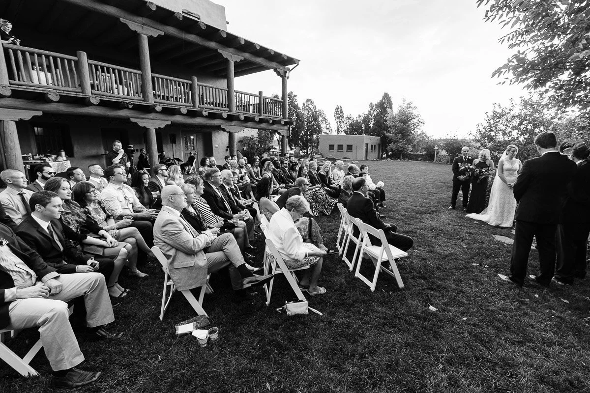 Outdoor wedding scene in black and white. Guests sit on chairs facing a couple and officiant. A rustic building and trees form the backdrop.