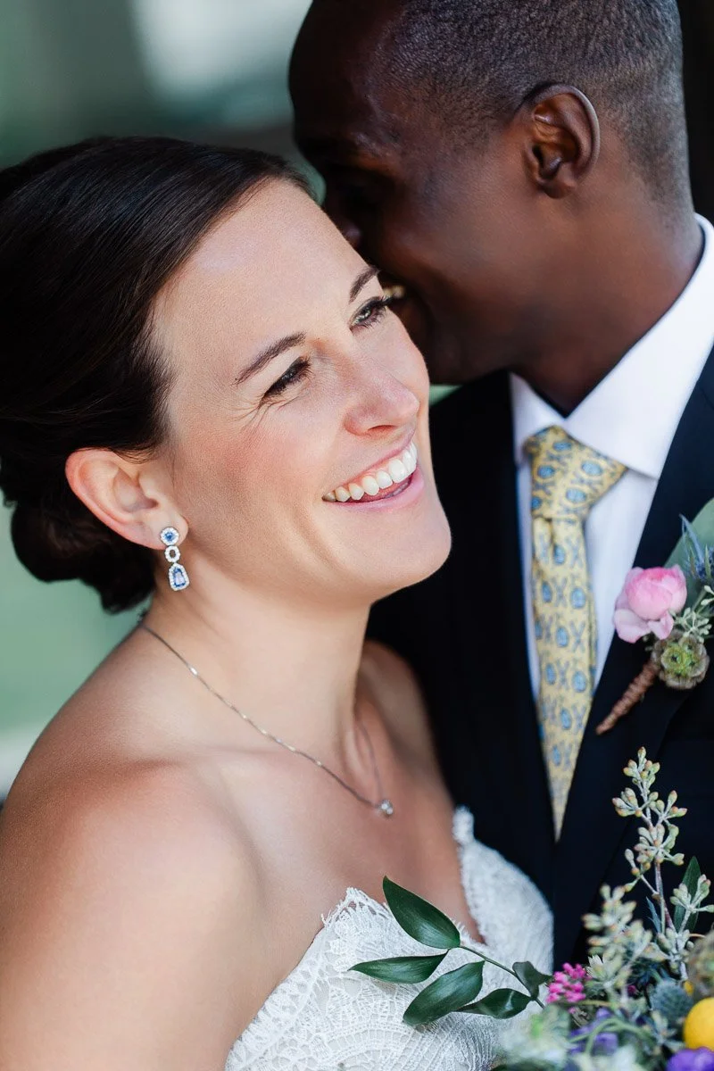 A joyful bride, in a lace wedding dress and blue earrings, smiles brightly while holding flowers. The groom whispers, creating a warm, intimate moment.