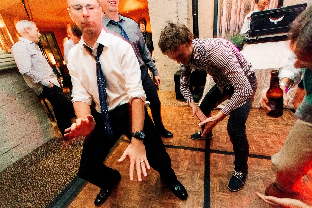 Group of men dancing energetically at a lively indoor party. Two men in shirts and ties are crouched, smiling, creating a joyful and carefree atmosphere.