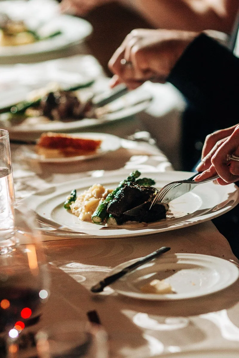 Close-up of a person cutting steak on a white plate with mashed potatoes and asparagus, set on a dining table. The scene conveys an elegant dining atmosphere.