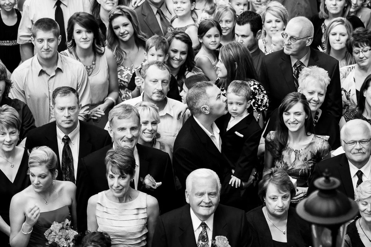A black and white image shows a large group at a formal event. In the center, a man kisses a child's cheek. The crowd appears joyful and celebratory.
