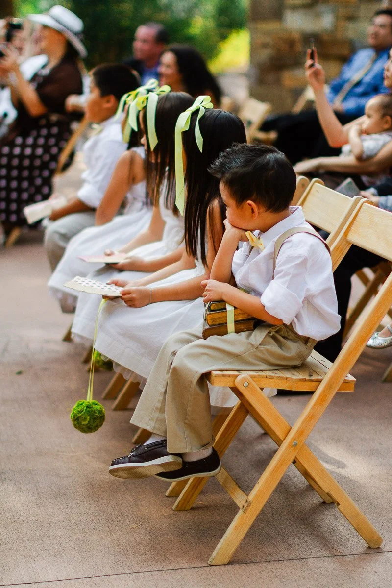Children seated in a row of wooden chairs at an outdoor event. They wear formal attire with the girls adorned with green ribbons, creating a festive atmosphere.