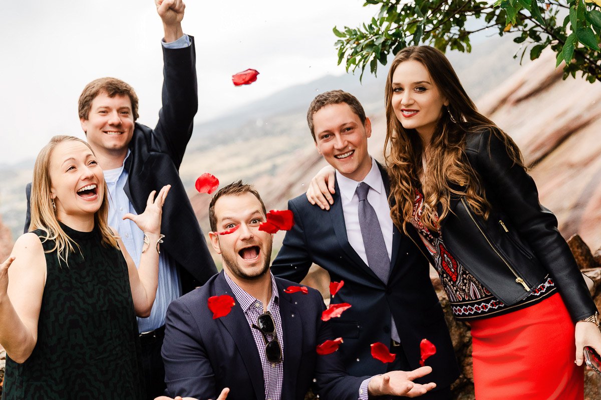 A group of five joyful people dressed in formal and casual attire, surrounded by falling red rose petals. Mountains and greenery in the background enhance the celebratory mood.