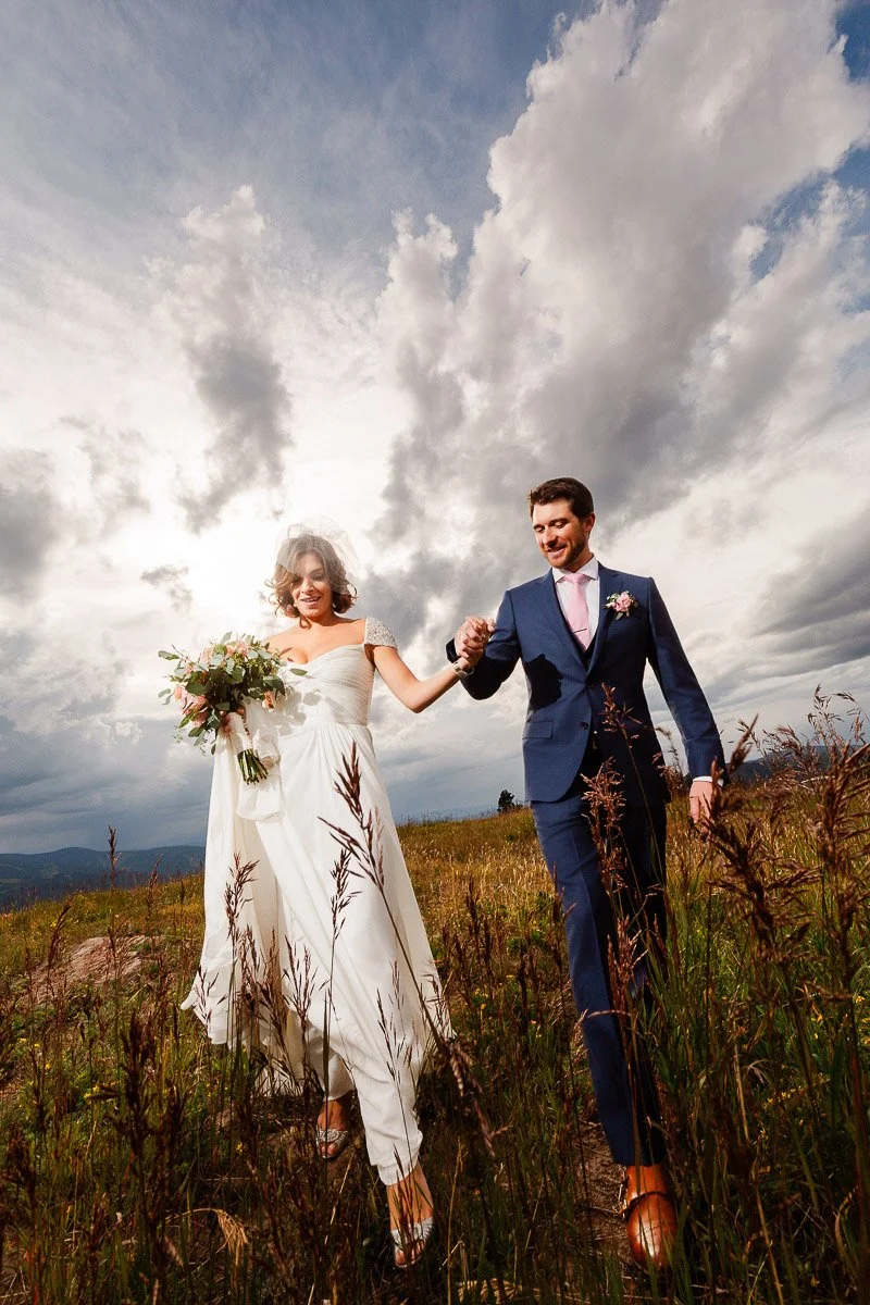 Bride and groom joyfully walk hand in hand through a grassy field. She holds a bouquet, and he wears a blue suit. Dramatic clouds fill the sky.