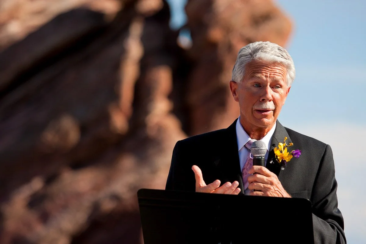 Elderly man in a suit speaks, holding a microphone, against a backdrop of red rock formations. He wears a serious expression, with a boutonniere adding color.