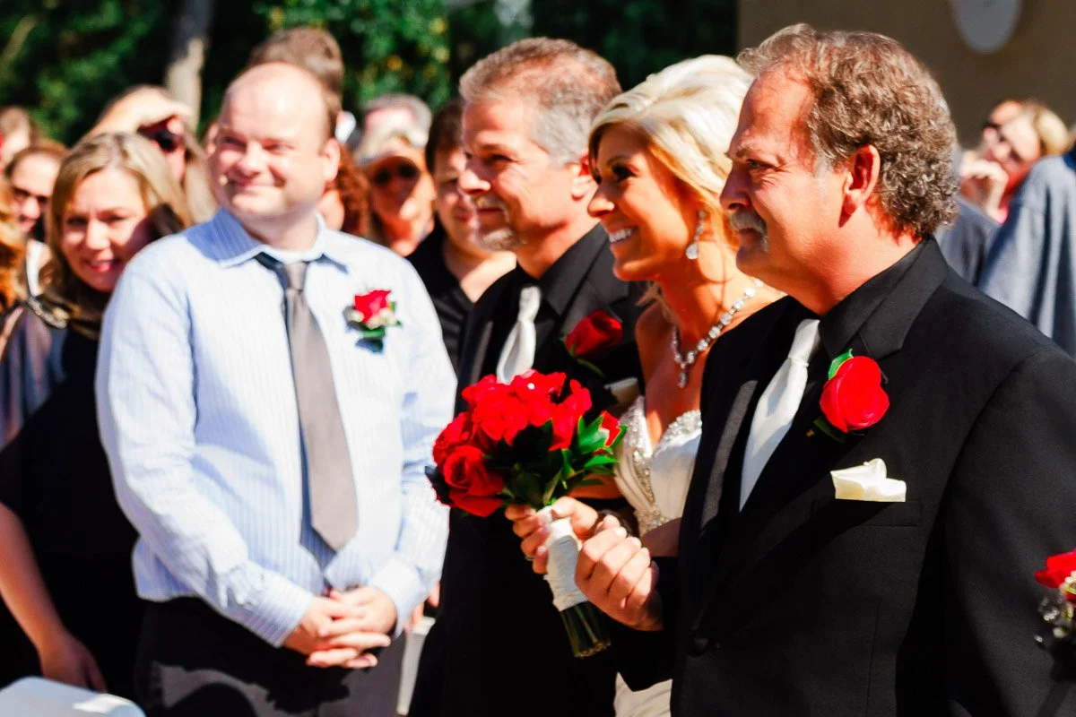 A smiling bride holding a bouquet of red roses walks arm-in-arm with two men in black suits at an outdoor wedding, surrounded by cheerful guests.
