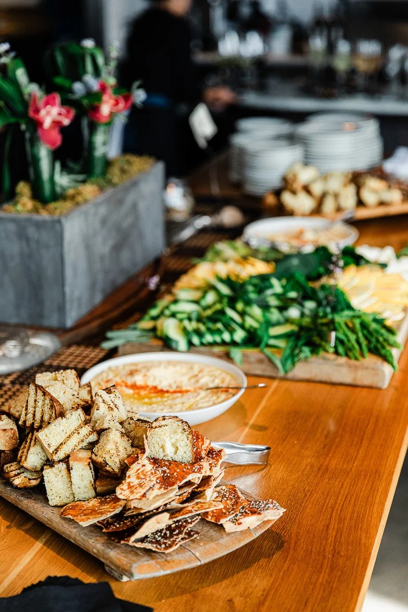 A wooden table with a buffet spread featuring sliced bread, crackers, hummus, mixed vegetables, and decorative flowers. The setting feels welcoming and appetizing.