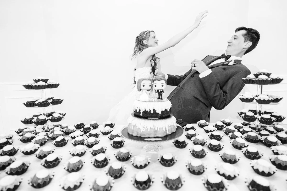 A bride playfully leans toward the groom, who reacts with surprise. They're surrounded by a table of cupcakes and a cake, suggesting a joyful wedding atmosphere.