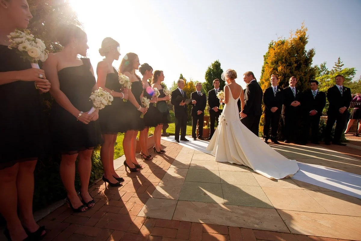 Outdoor wedding ceremony with bridesmaids in black dresses and groomsmen in suits. The bride and groom stand under bright, warm sunlight during a York Street Botanic Gardens wedding in Denver, Colorado