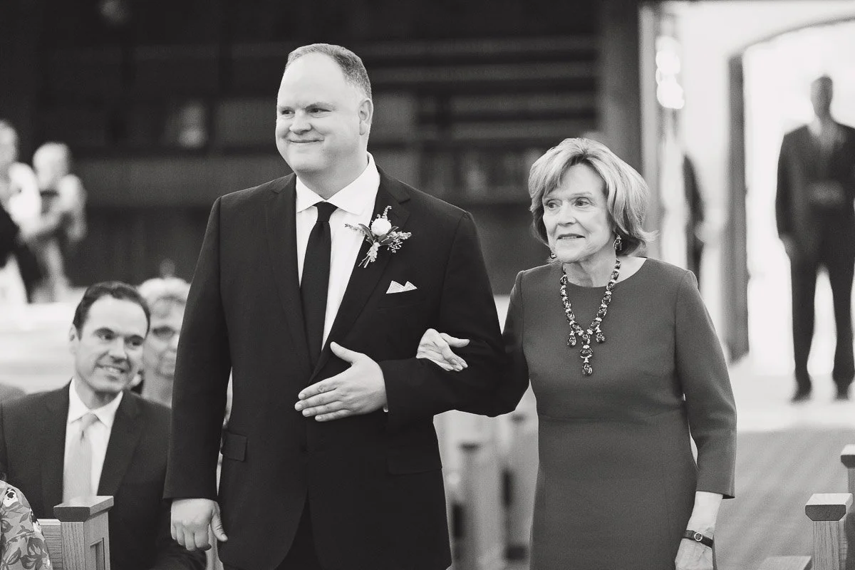 A man in a suit with a floral boutonnière escorts an older woman in a dress holding his arm down an aisle. Both appear happy and composed in a formal setting.
