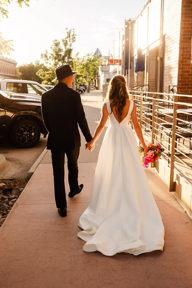 A couple walks hand in hand away from the camera on a sunlit street. The bride wears a white gown, holding a bouquet, while the groom is in a black suit and hat.