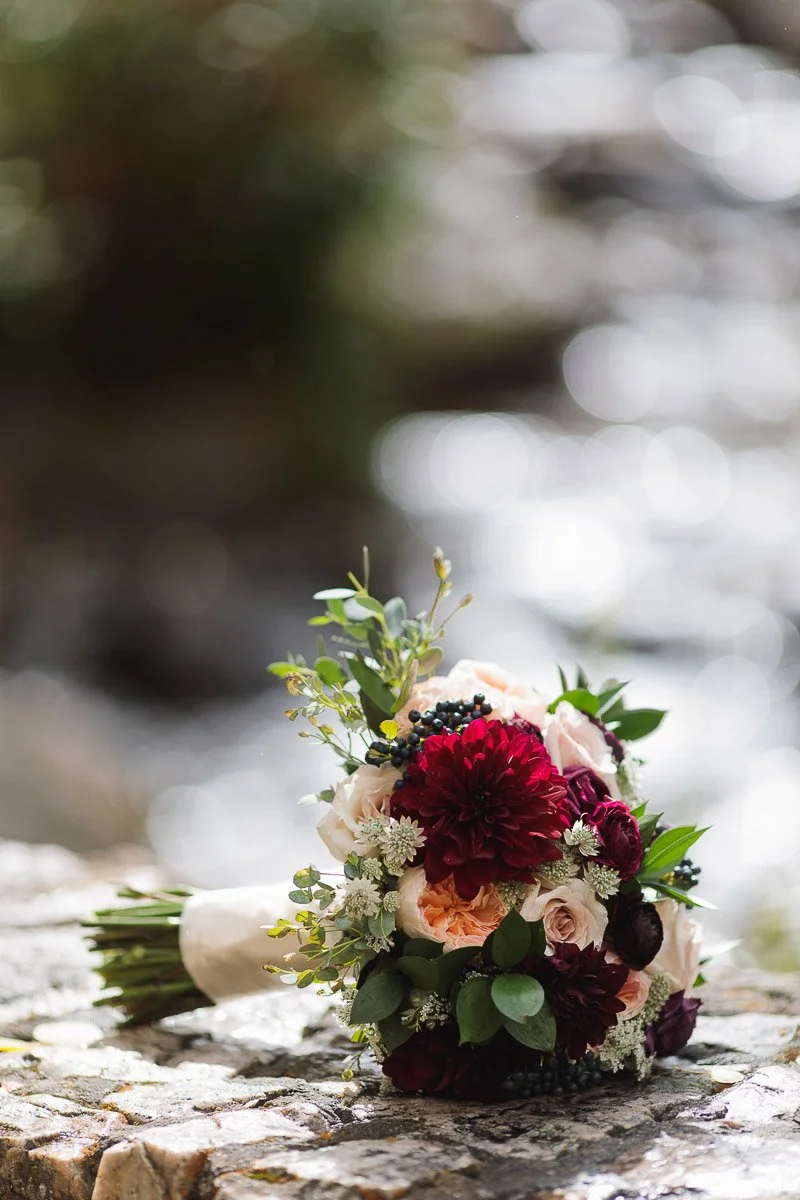 A vibrant bouquet of burgundy and peach flowers with greenery lies on a sunlit stone surface. The blurred background suggests a serene, natural setting.