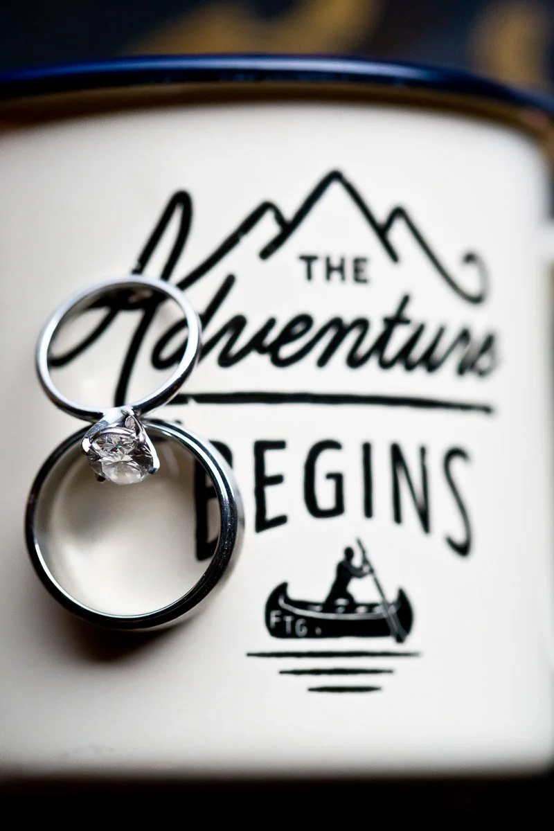 Two rings, one with a diamond, rest against a mug reading "The Adventure Begins." A design of mountains and a canoe adds a sense of journey and new beginnings.