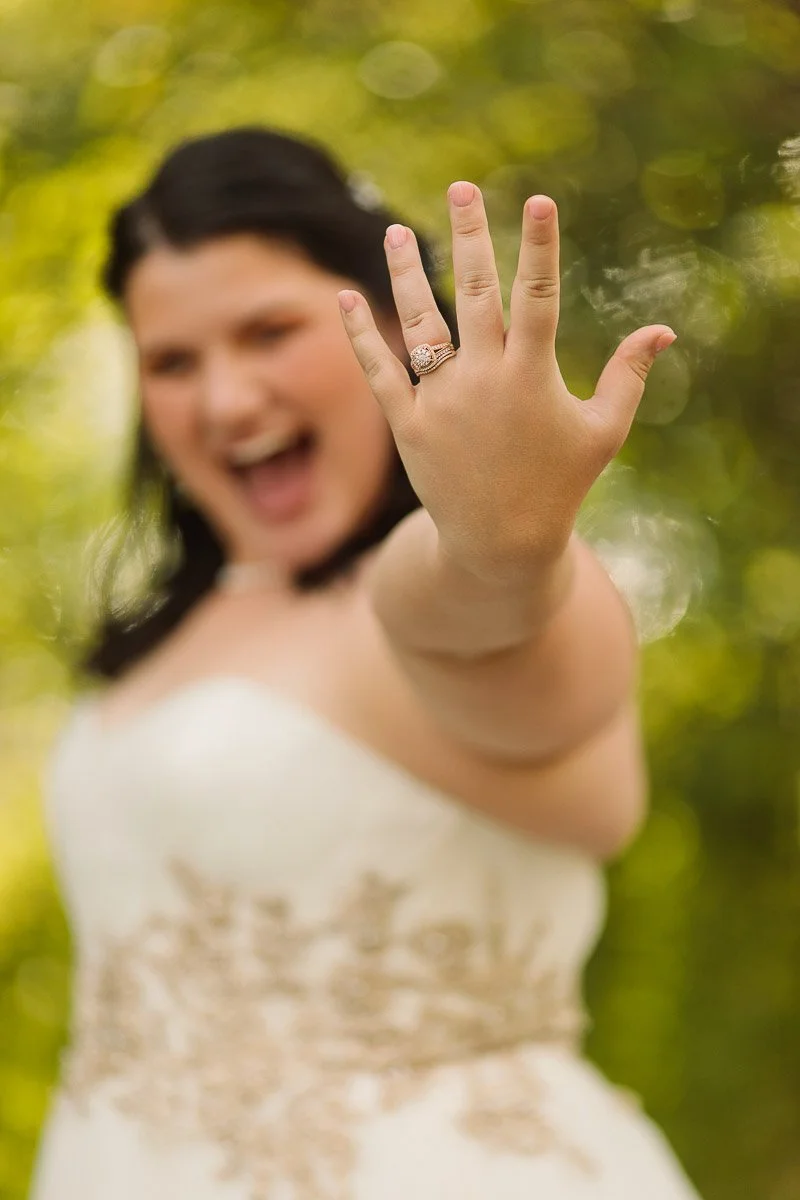 A joyful bride in a white gown displays her engagement ring prominently, her face blurred in the background, amidst a sunny green backdrop.