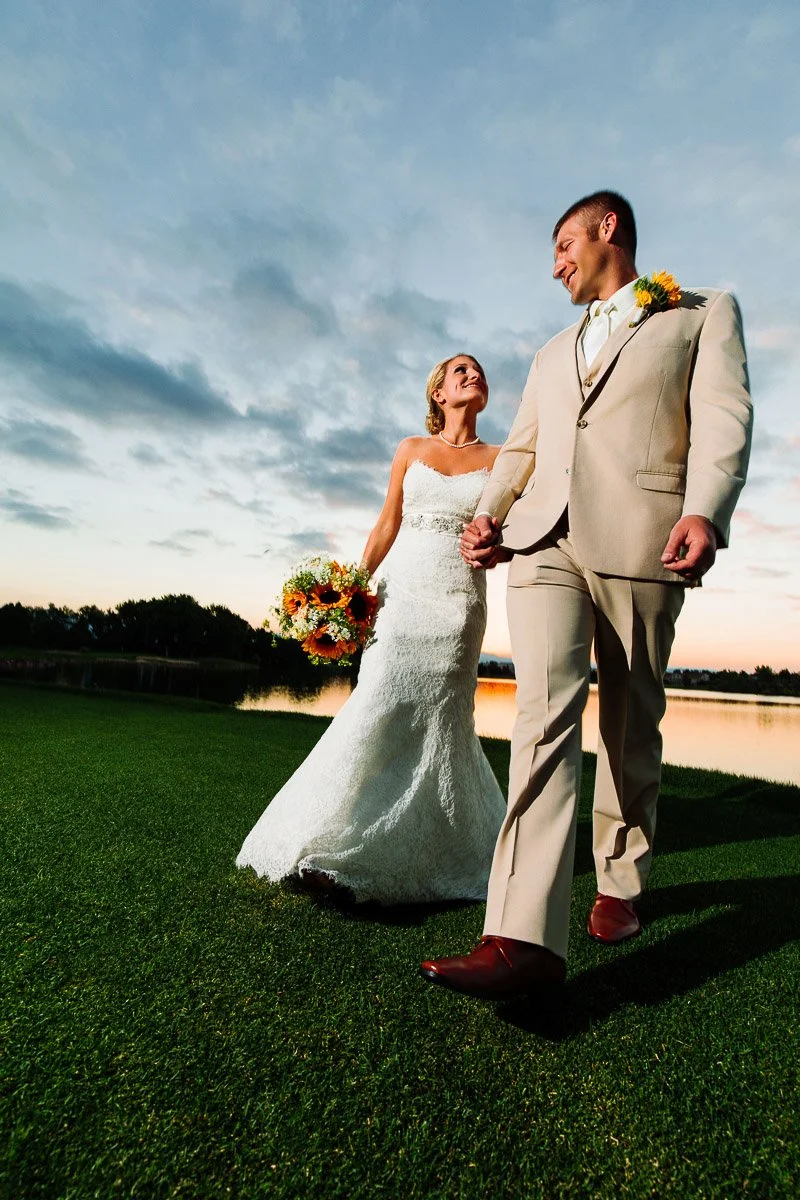 Bride and groom holding hands, walking on grass at sunset. She wears a white lace dress, he a beige suit. Both smiling, with a serene lake backdrop.