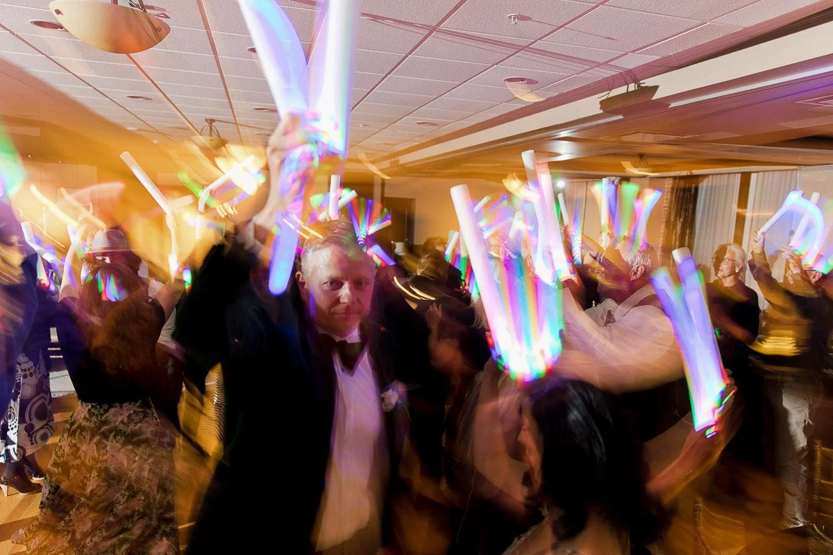 A lively party scene with a crowded dance floor. Guests are enthusiastically waving colorful, glowing sticks, creating a vibrant, energetic atmosphere.
