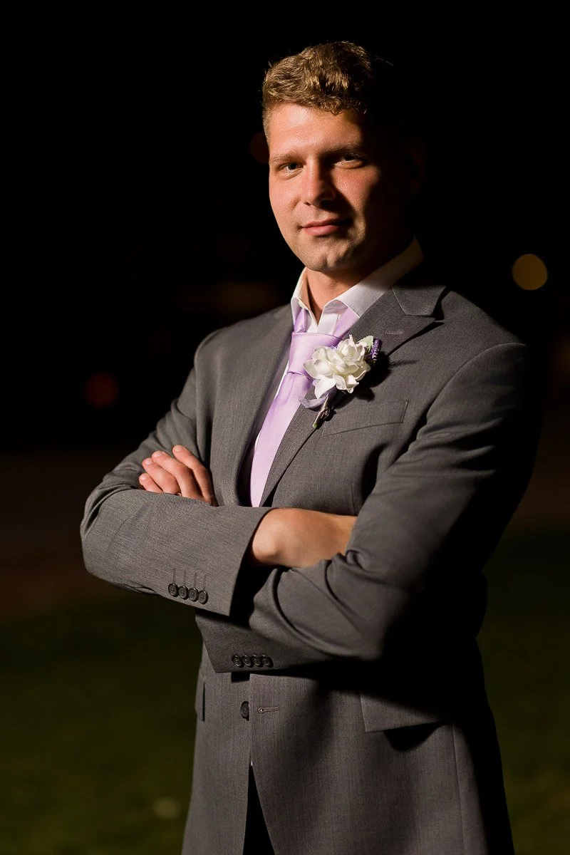 Man in a gray suit with a lavender tie stands confidently with crossed arms at night. He has a white floral boutonniere and a serene expression.