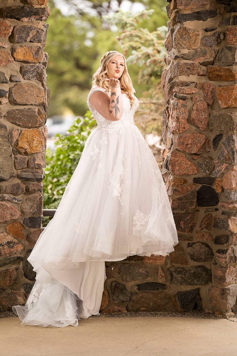 Bride in a flowing white gown stands between stone pillars, surrounded by greenery. She smiles softly, exuding a serene and joyful mood.
