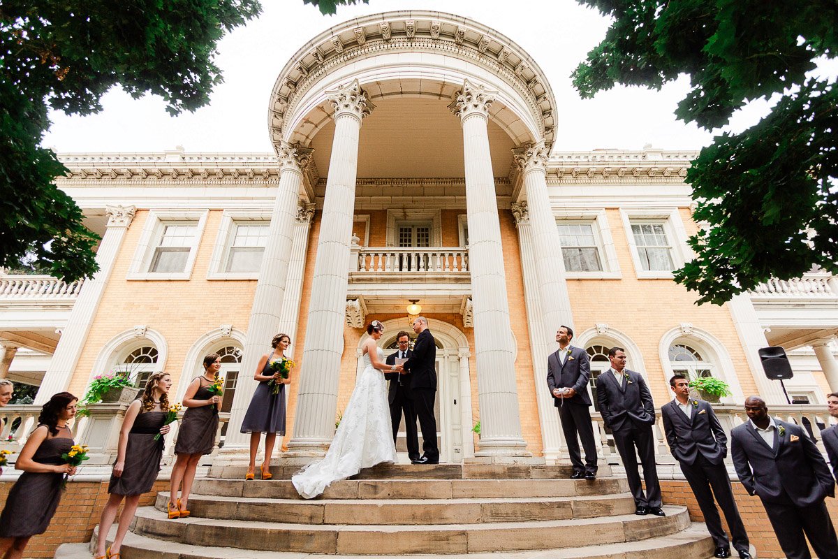 A wedding ceremony takes place on the steps of a grand mansion with tall columns. The bride and groom stand at the center, surrounded by an elegant bridal party.