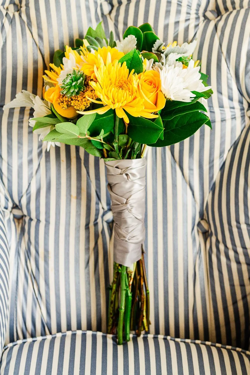 A vibrant bouquet featuring yellow roses, chrysanthemums, and daisies with lush green foliage, wrapped in silver ribbon, rests on a striped chair.