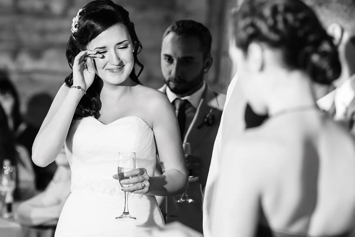 A bride in a strapless gown wipes tears while holding a champagne glass, surrounded by guests in a warmly lit setting, conveying joy and emotion.