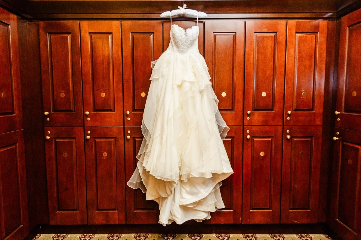 Elegant ivory wedding gown with ruffled layers hangs against a backdrop of rich wooden lockers, creating a warm, sophisticated ambiance.