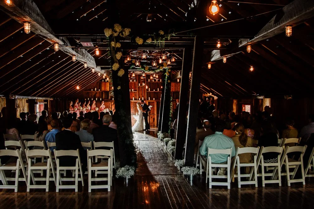 A rustic wedding ceremony in a dimly lit barn with wooden beams. A couple stands at the altar, surrounded by white flowers and seated guests.