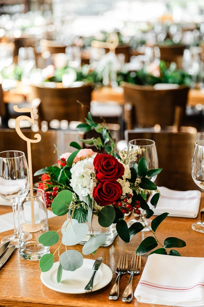 Elegant table setting with a centerpiece of red and white roses, eucalyptus, and berries. Glasses, cutlery, and napkins add a formal touch.
