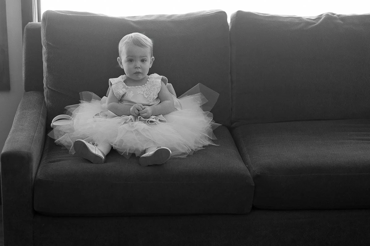 A young child in a tutu dress sits on a sofa, gazing calmly at the camera. The black-and-white image conveys a serene, elegant atmosphere.