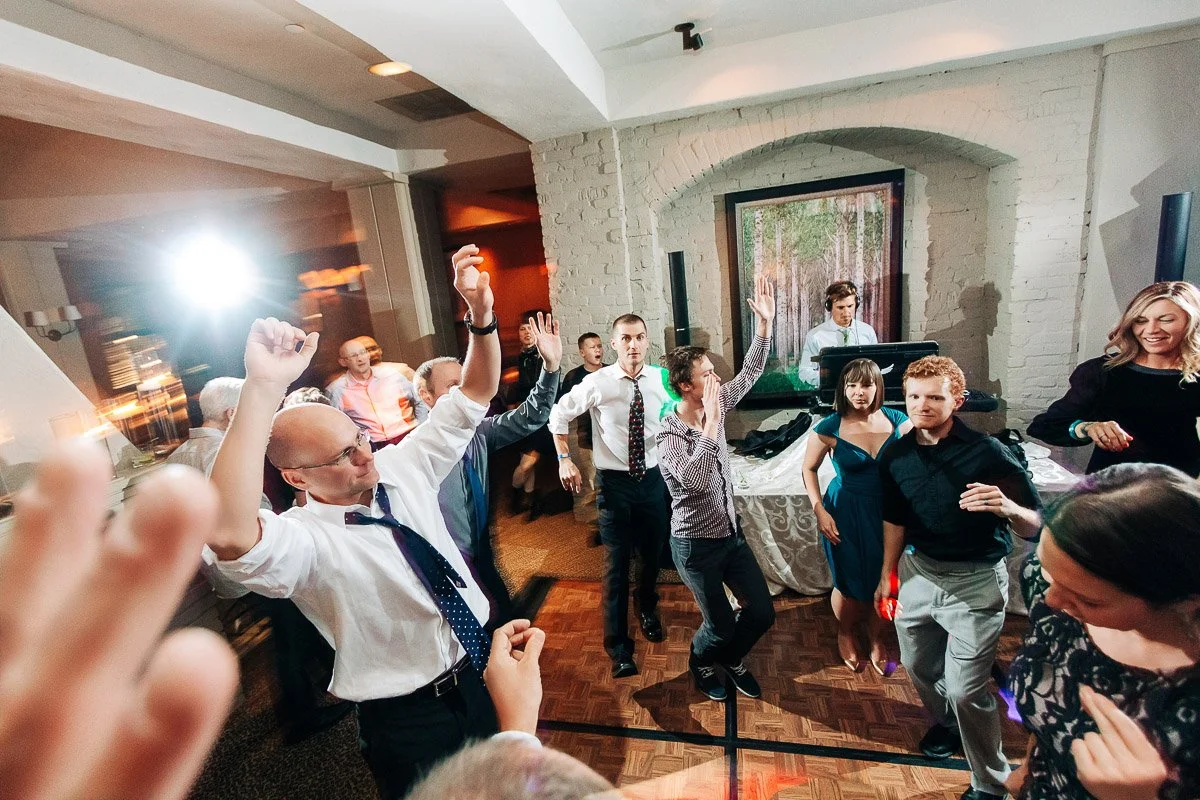 A lively party scene with people dancing energetically inside a room with white walls and wooden floors. A DJ stands near the back, playing music.