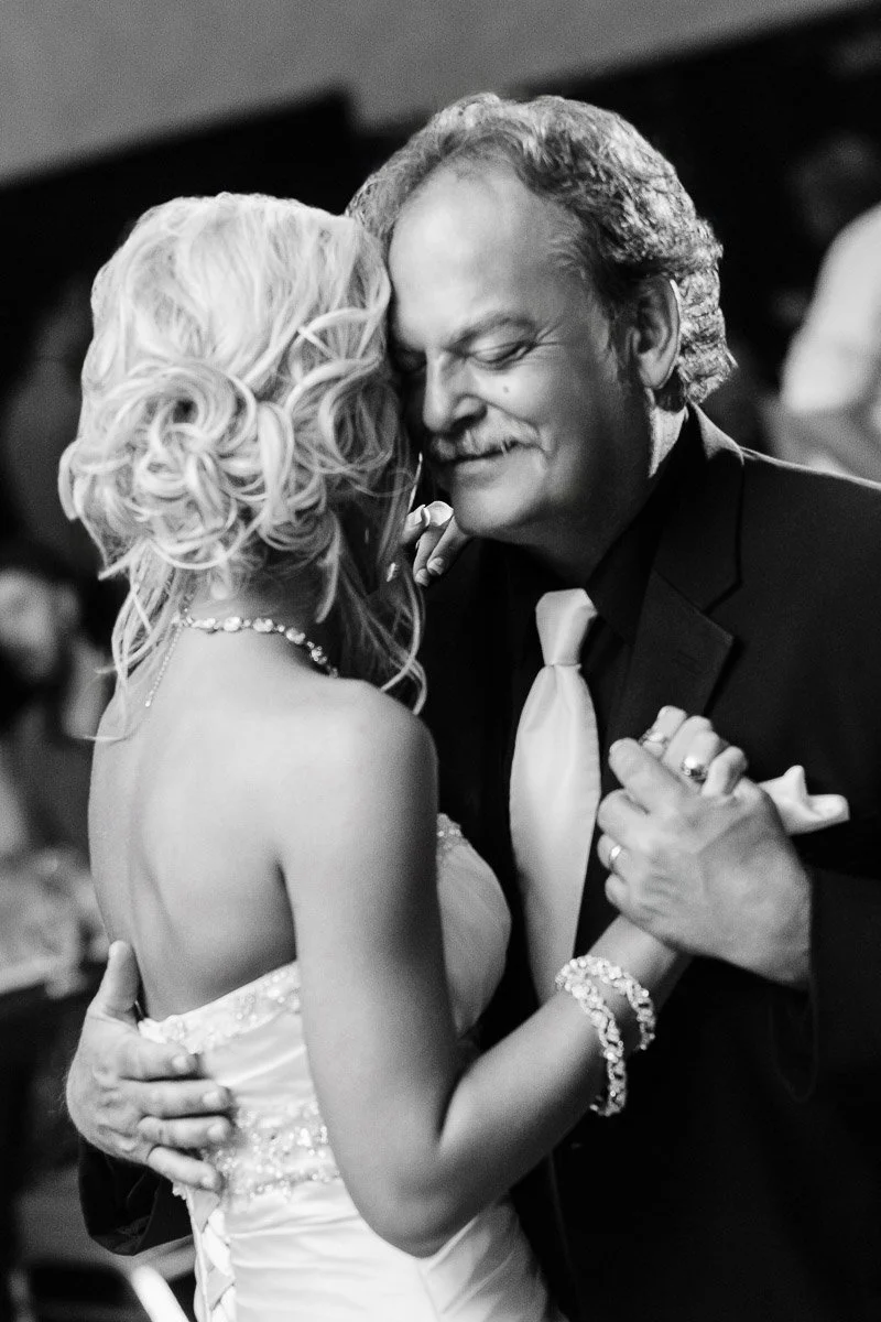 A black and white photo of a joyful couple dancing closely. The woman in a wedding dress and the man in a suit share a tender, smiling embrace.