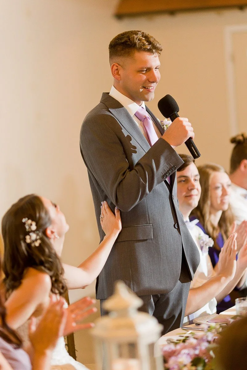 A man in a gray suit with a purple tie is smiling while speaking into a microphone at a wedding. A woman, seated nearby, touches his arm affectionately.