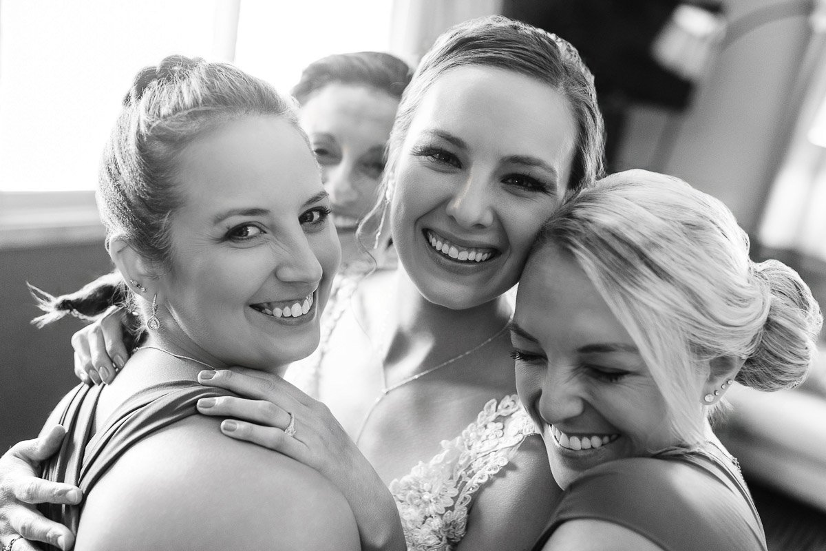 Black and white photo of four women smiling and embracing, conveying joy and closeness. The bride is in the center, highlighted by soft lighting.