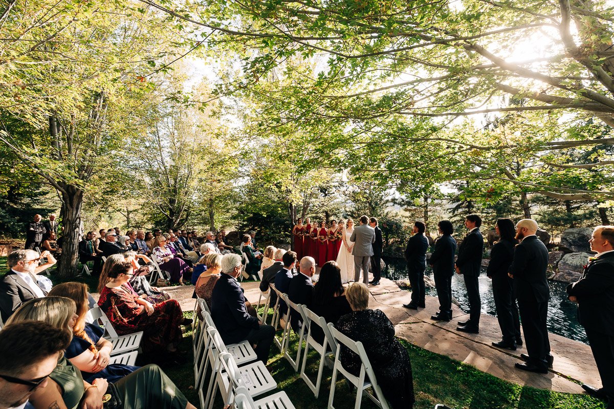 Outdoor wedding ceremony under sunlight with lush trees. Guests seated on white chairs face the couple and wedding party by a serene pond.
