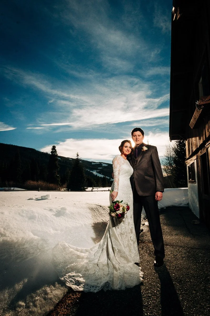 Bride in a lace gown and groom in a suit stand on a snow-covered path by a wooden cabin under a vibrant blue sky with scattered clouds.